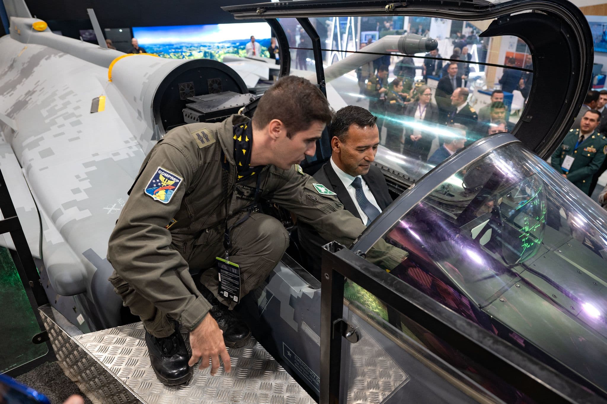 Pedro Sánchez Suárez, de traje, se sienta en la cabina de un avión de combate mientras un piloto le explica la instrumentación, durante una feria o exposición de defensa con público al fondo.