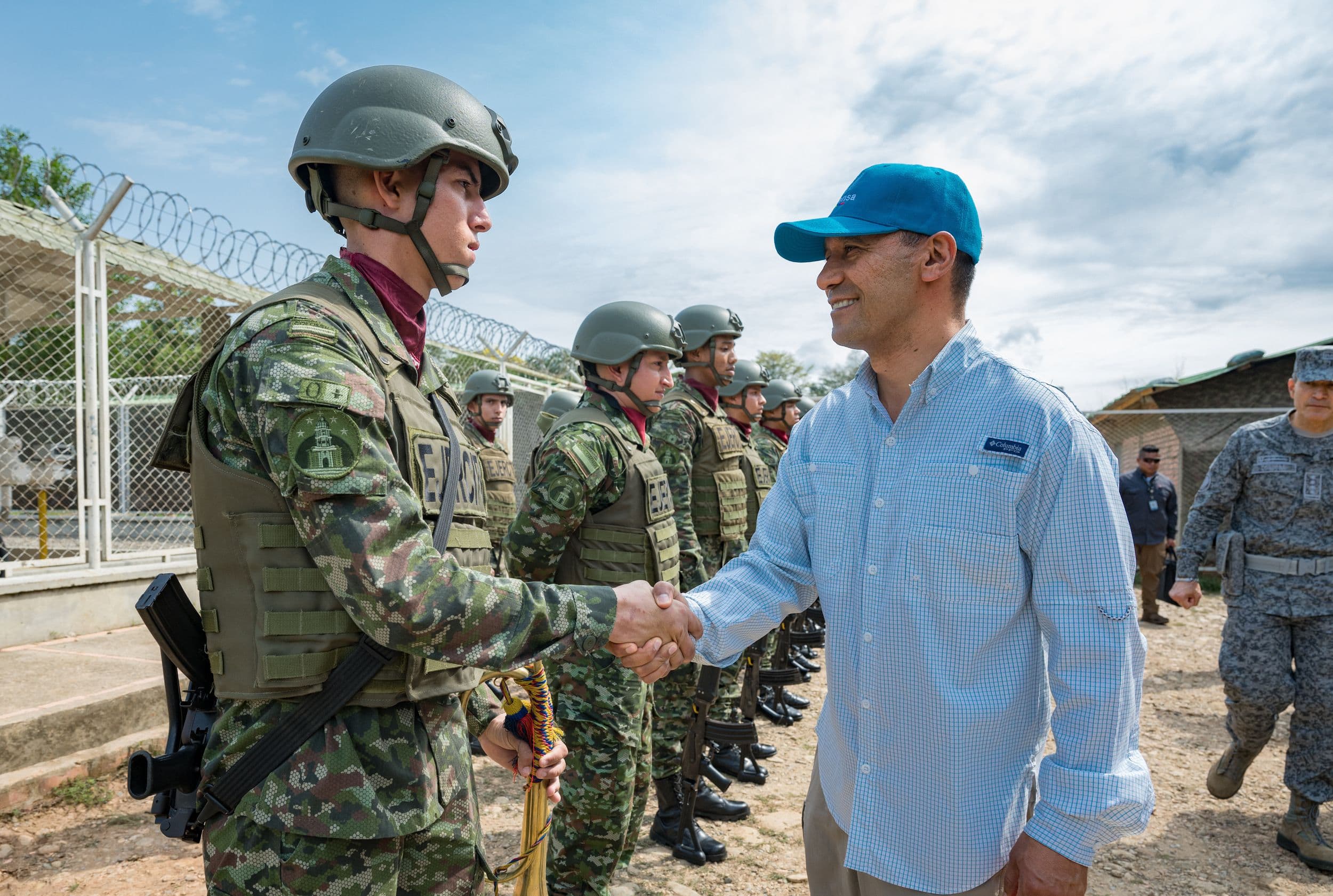 Pedro Sánchez Suárez, de civil, saluda de mano a un soldado con casco y equipo táctico, frente a una fila de militares formados junto a una malla con alambre de púas.