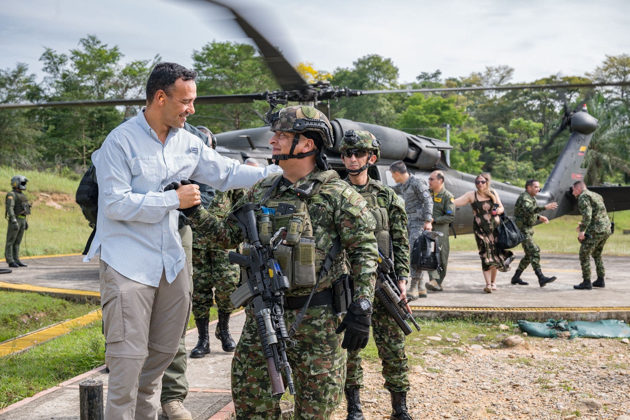 Pedro Arnulfo Sánchez Suárez, vestido de civil, saluda con la mano en el hombro a un soldado con casco y fusil, frente a un helicóptero militar en una pista, rodeados de más uniformados.