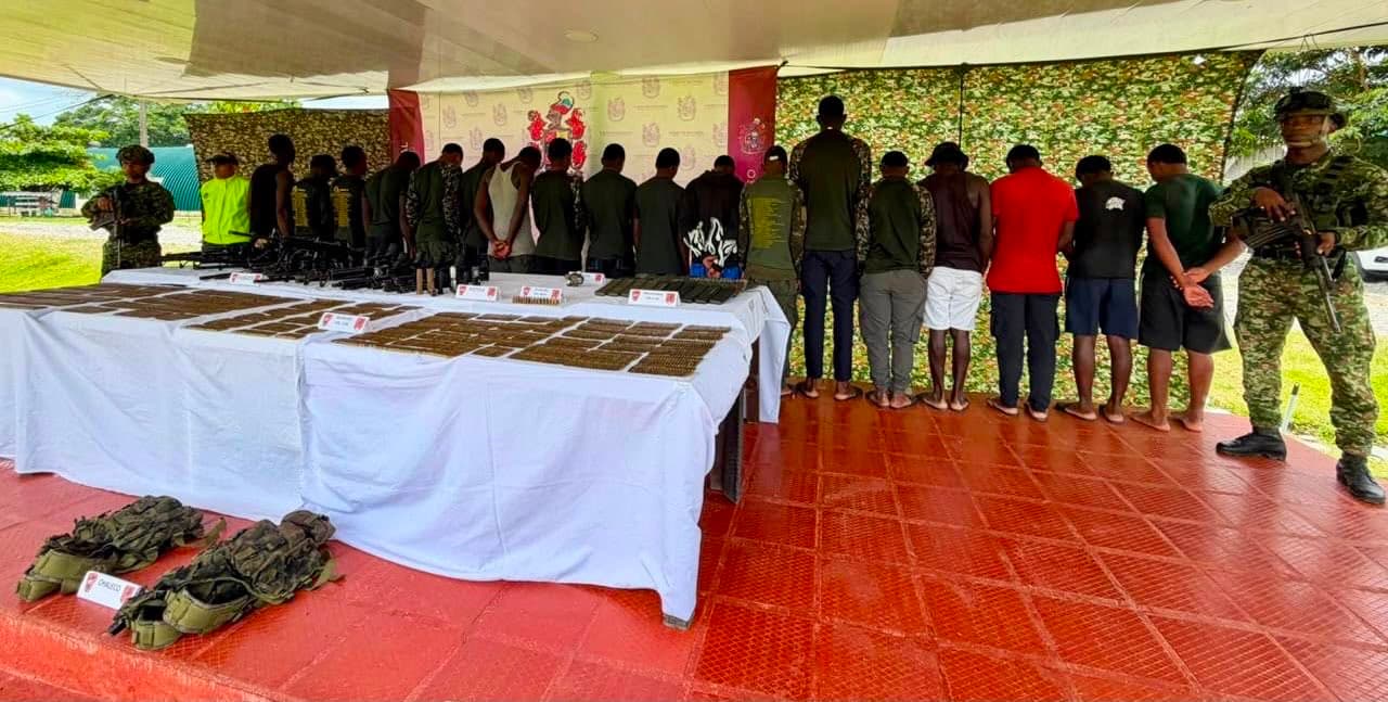 Un grupo de personas capturadas, de espaldas y con las manos esposadas, custodiadas por un militar armado, junto a mesas con armas, munición y equipo militar incautado, durante una presentación de resultados operativos.