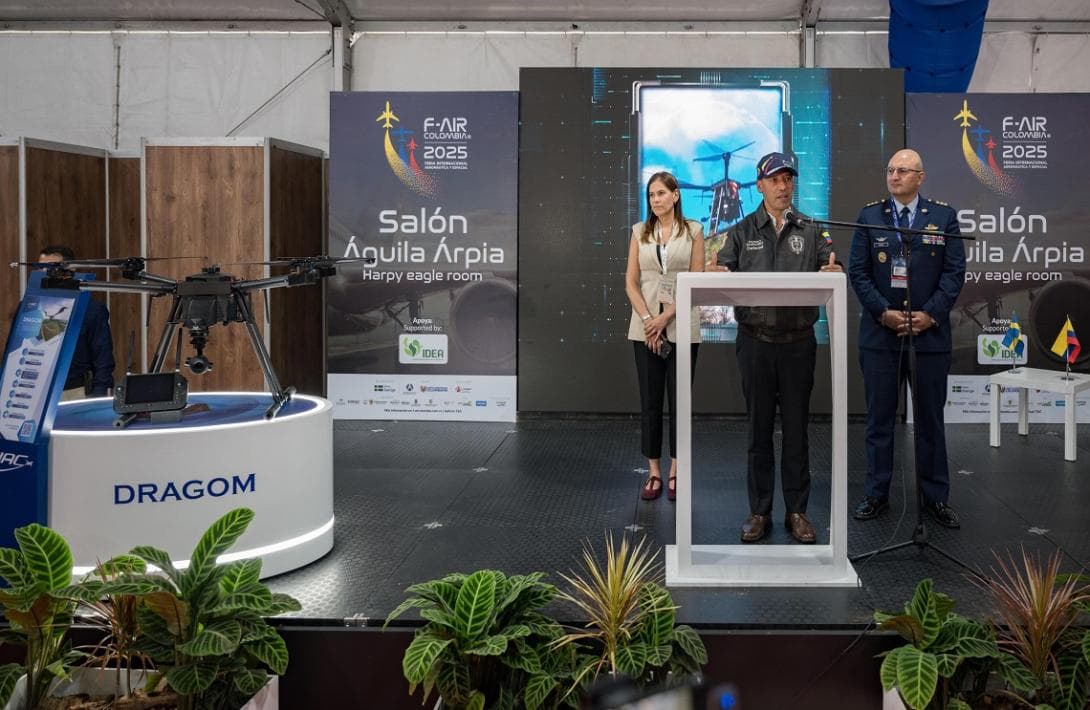 Pedro Sánchez Suárez habla en un atril con micrófono junto a otras dos personas en un escenario, con un dron exhibido en primer plano y afiches de "F-AIR Colombia 2025 - Salón Águila Árpia" al fondo.