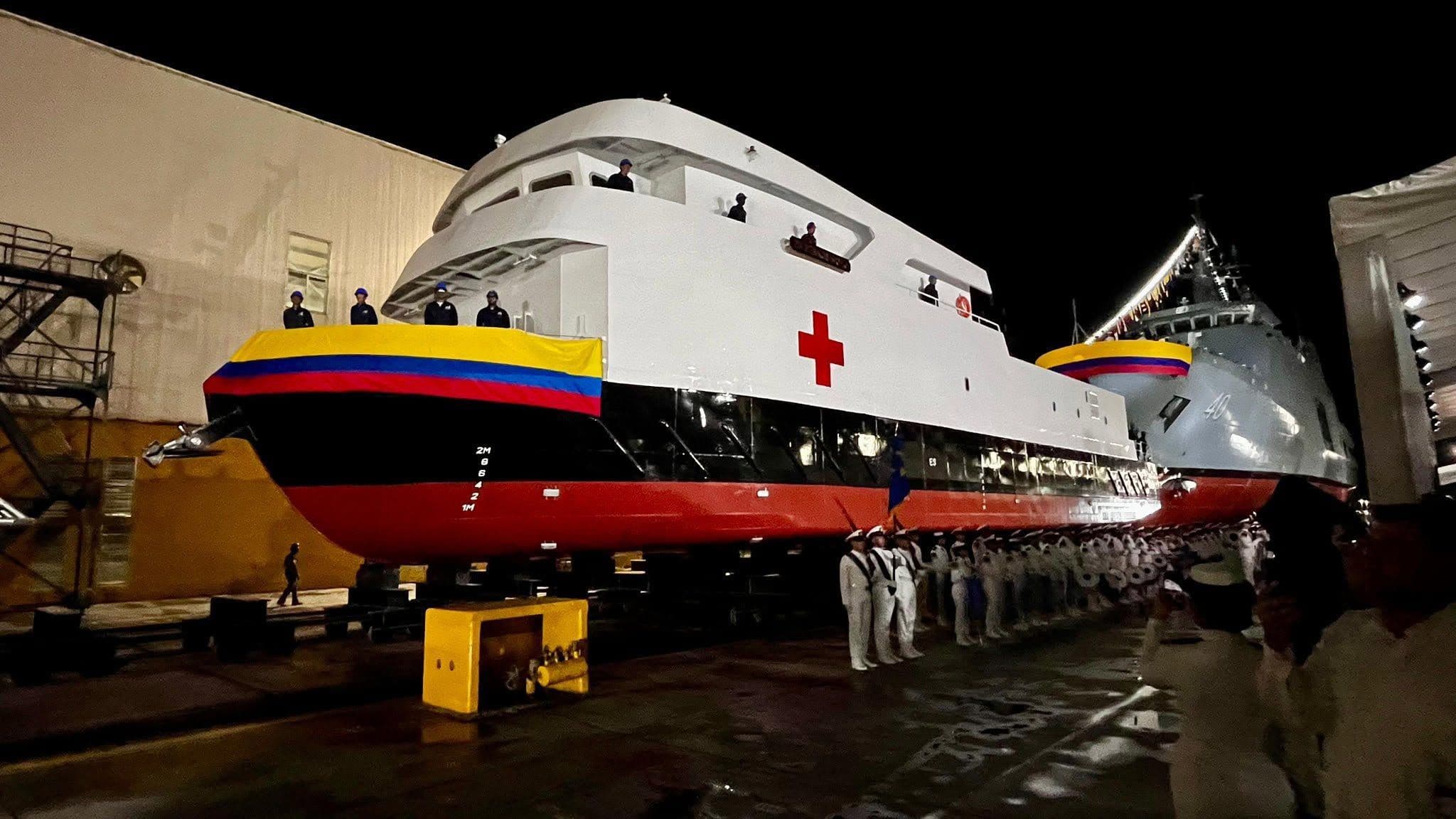 Buque hospital, con proa pintada con la bandera de Colombia y una cruz roja, sobre rieles en un astillero de noche, junto a una fila de marinos de uniforme blanco formados.