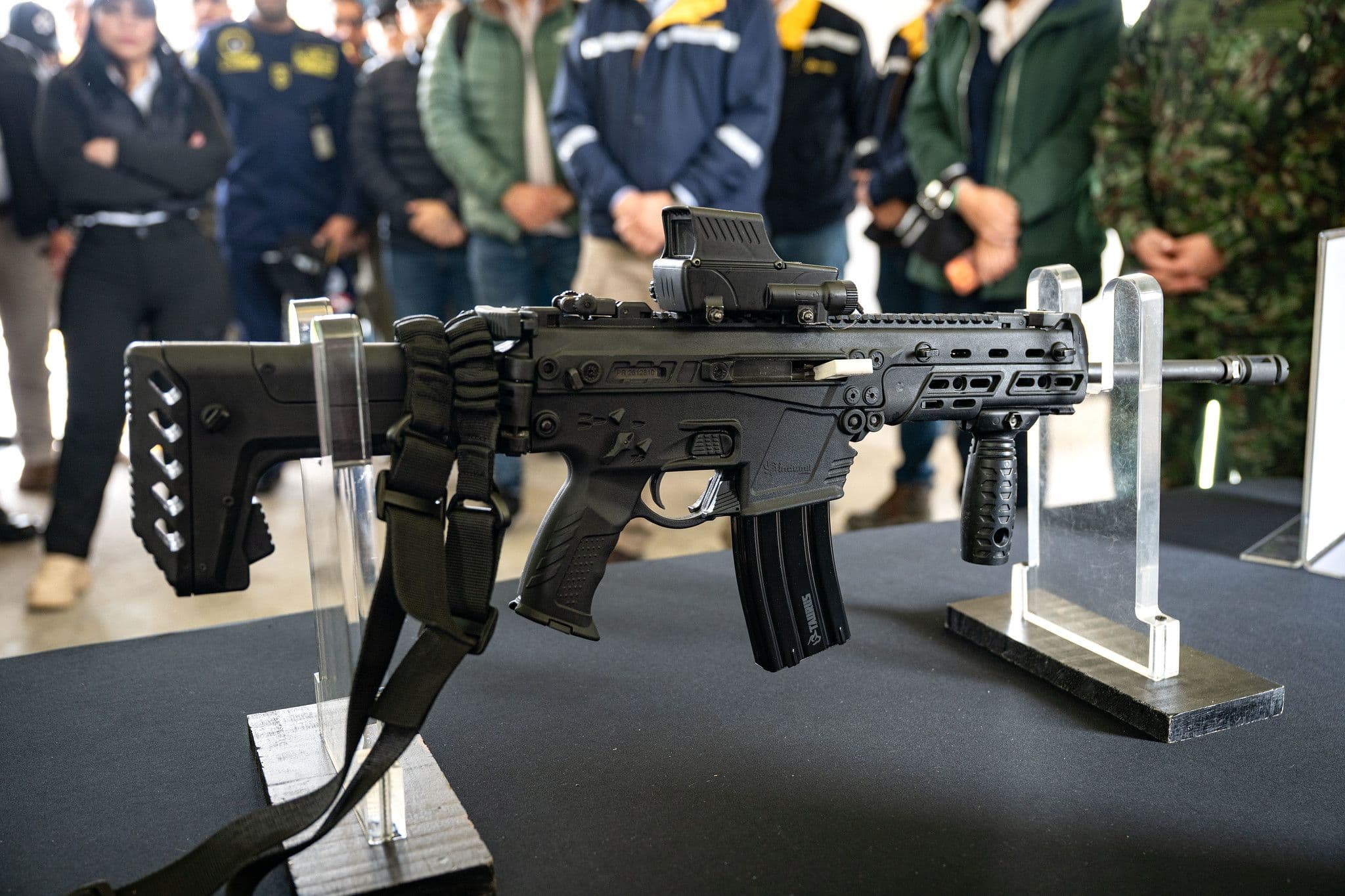 Fusil de asalto con mira holográfica exhibido sobre soportes en una mesa, rodeado de personas observando, durante una feria o exposición de industria de defensa.