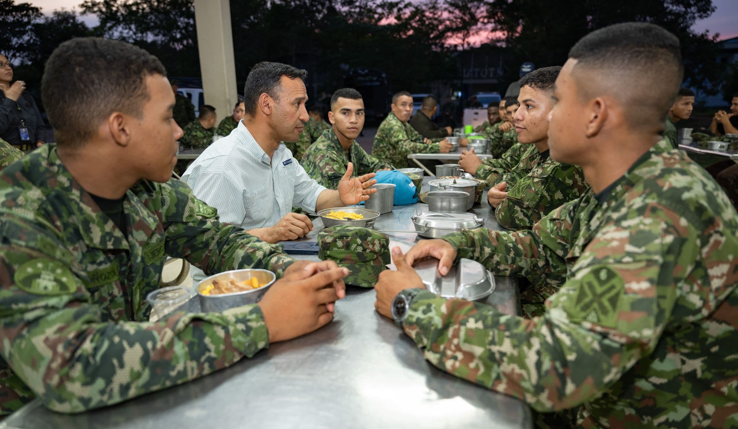 Pedro Sánchez Suárez, de civil, comparte una comida y conversa con soldados en una mesa metálica al aire libre, al atardecer.