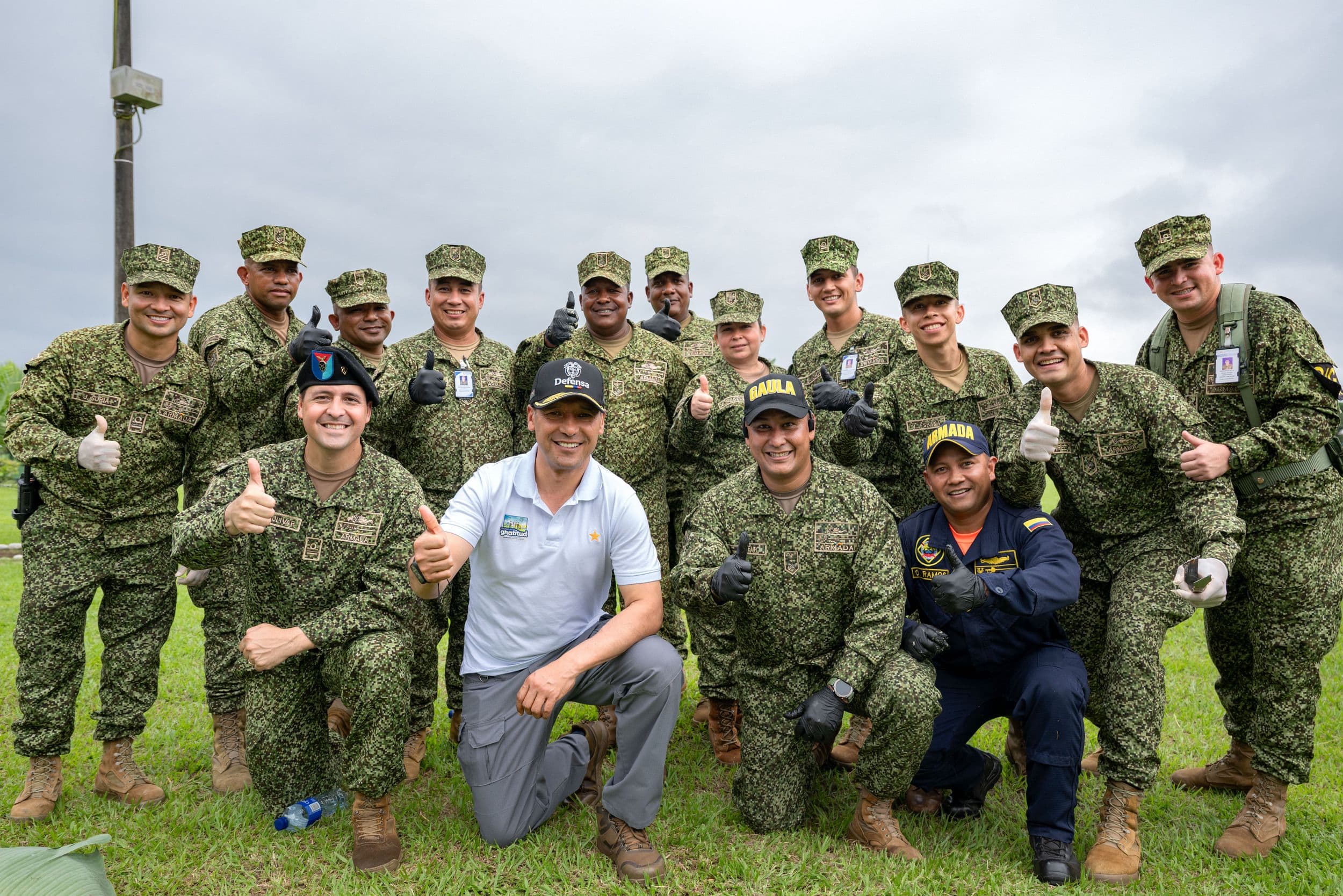 Pedro Sánchez Suárez posa arrodillado junto a un grupo de miembros de la Armada Nacional en uniforme de campaña, la mayoría con el pulgar arriba, en una foto grupal al aire libre sobre césped.