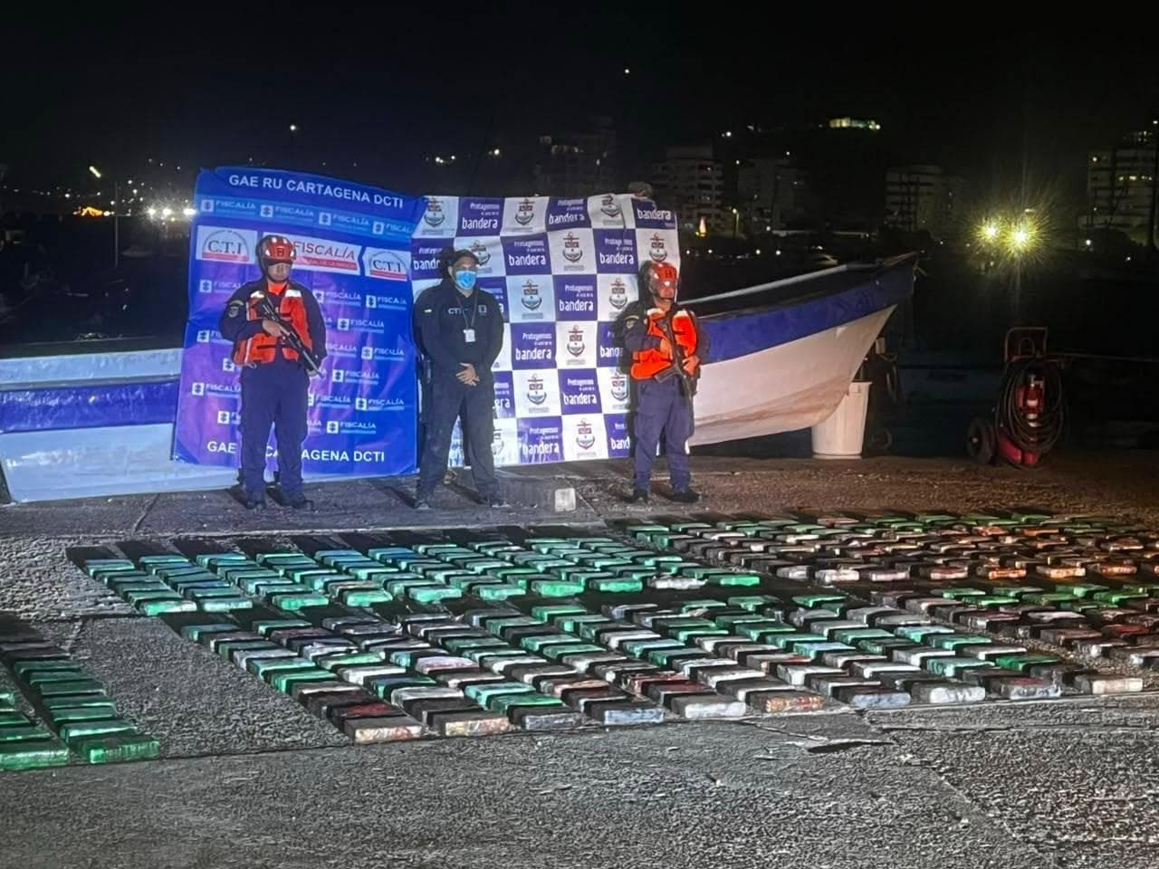 Filas de paquetes envueltos, presuntamente droga incautada, exhibidos en un muelle de noche, con dos uniformados armados y un funcionario de la Fiscalía junto a una lancha y un pendón del CTI-GAE Ru Cartagena.