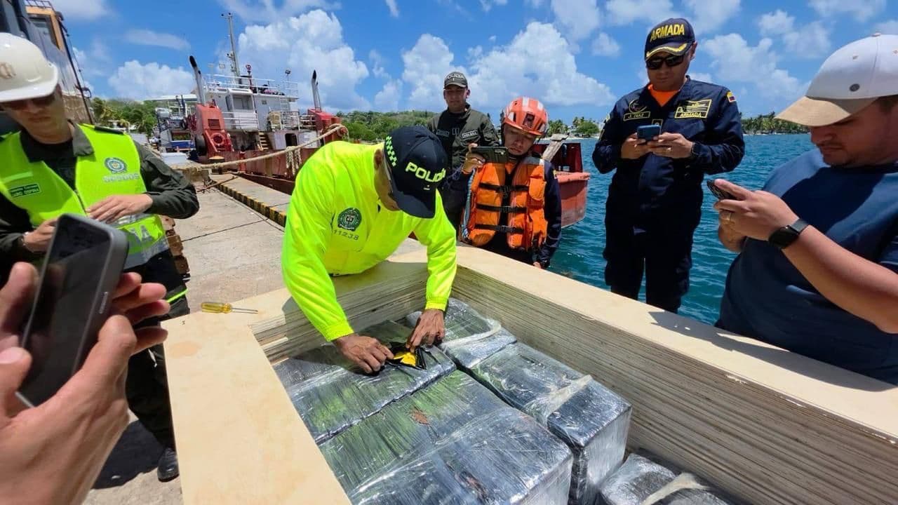 Un funcionario de la Policía inspecciona paquetes envueltos, presuntamente droga incautada, dentro de un contenedor de madera en un muelle, rodeado de personal de la Armada y personas grabando con sus celulares.