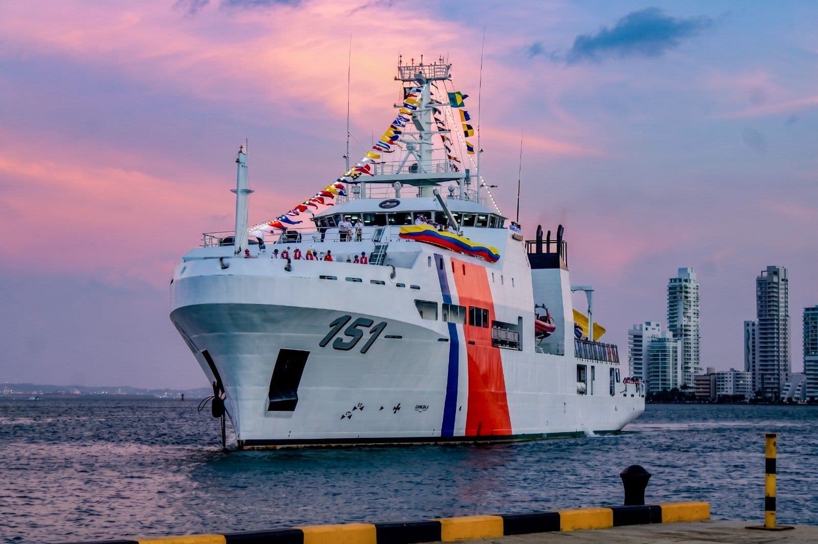 Buque de la Guardia Costera identificado con el número 151, engalanado con banderines, ingresando a la bahía de Cartagena al atardecer, con el perfil de edificios de la ciudad al fondo.