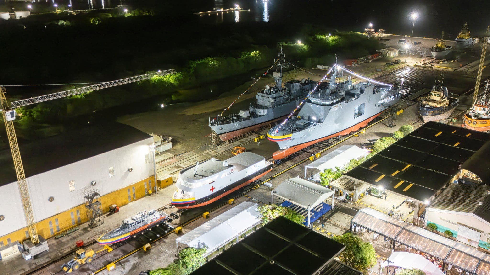 Vista aérea nocturna de un astillero con varios buques en construcción sobre rieles, incluyendo un buque hospital con cruz roja y un buque de guerra gris engalanado con banderines, rodeados de grúas y carpas para un evento.