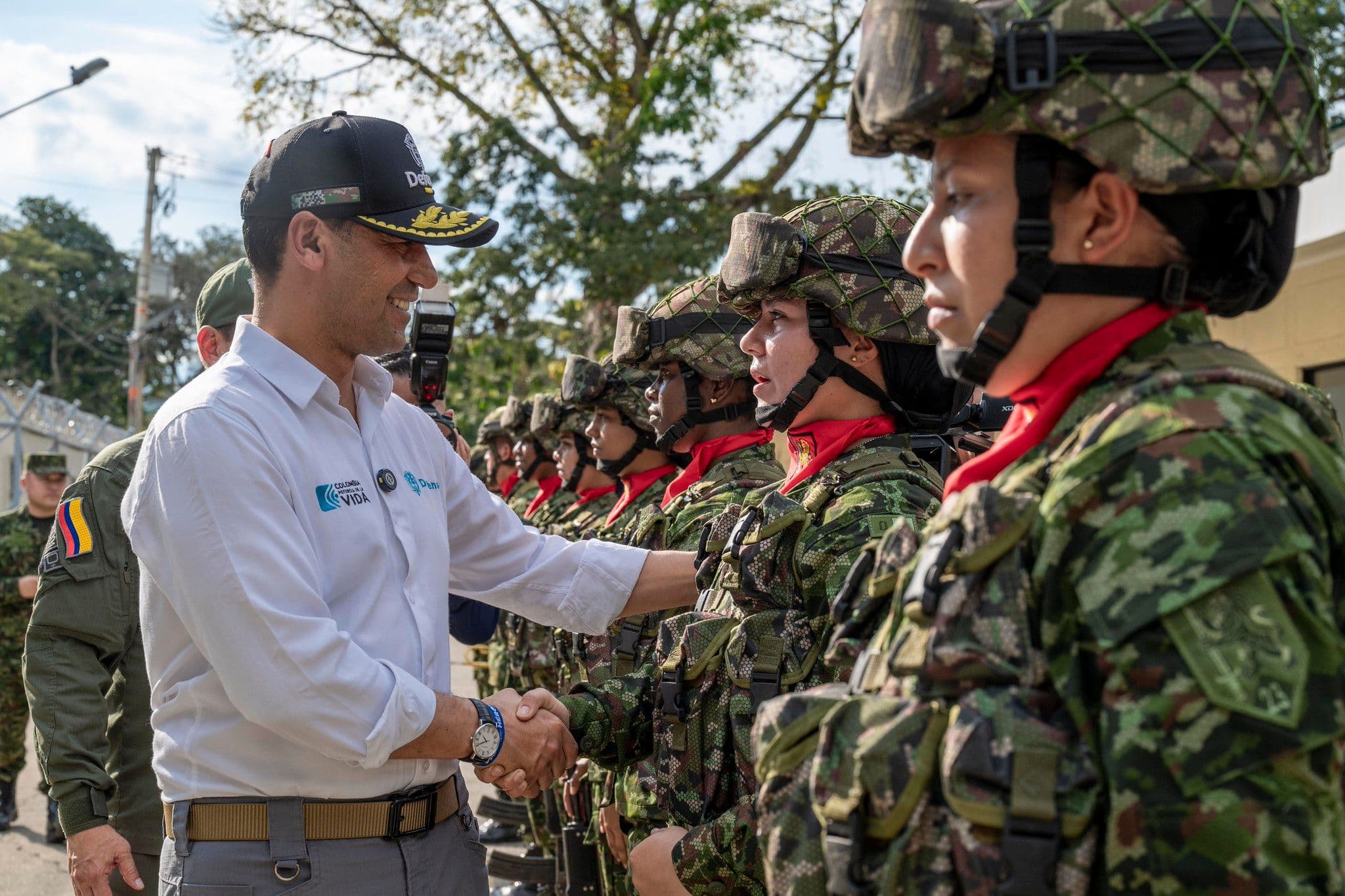 Pedro Sánchez Suárez, de civil, saluda de mano a una soldado con casco y equipo táctico, frente a una fila de militares en formación al aire libre.