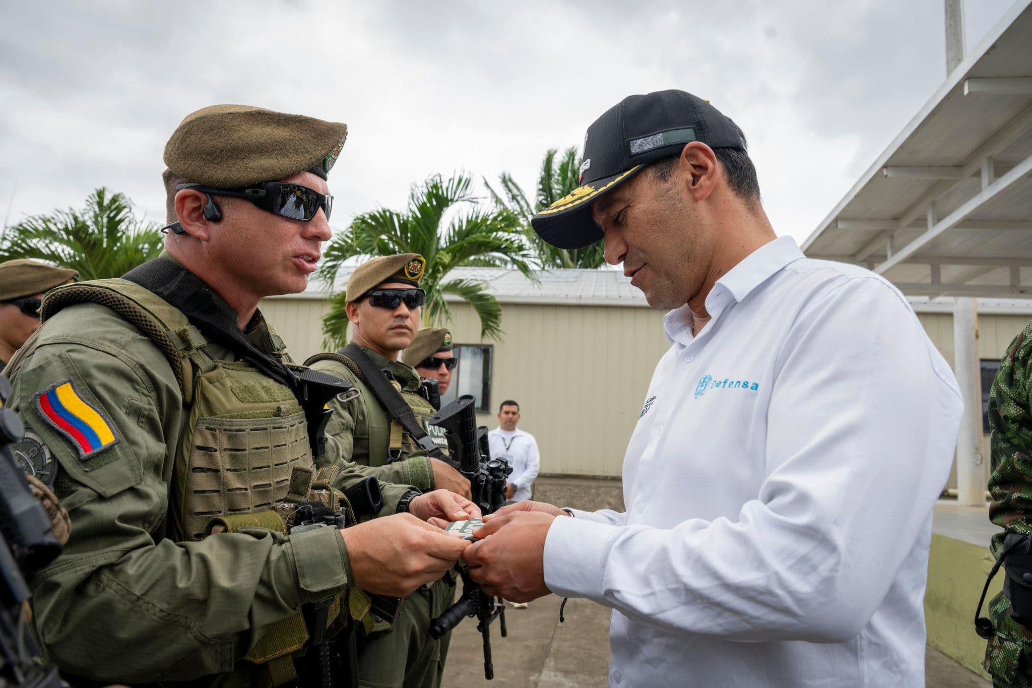 Una toma de plano medio muestra a Pedro Sánchez Suárez, vistiendo una camisa blanca de manga larga con el logo "Defensa" y una gorra negra con visera amarilla, recibiendo un objeto pequeño de un soldado uniformado. El soldado, de perfil, usa un uniforme verde militar con equipo táctico, boina marrón, y gafas de sol, mostrando la bandera de Colombia en su manga. Varios otros militares están de pie en el fondo, algunos armados con rifles. El fondo es un patio exterior con edificios y palmeras bajo un cielo nublado.