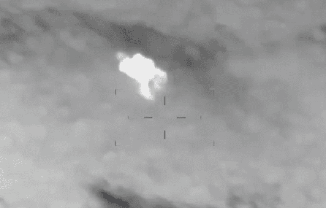 Imagen en escala de grises de un video de vigilancia aérea con visión térmica, mostrando un destello blanco de impacto y una mira reticular superpuesta sobre el terreno