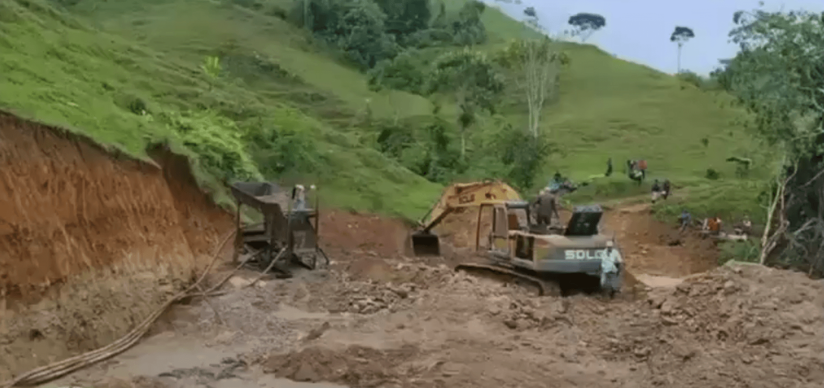 Excavadora y maquinaria de minería ilegal operando en un terreno de suelo removido junto a una colina verde, con varias personas observando al fondo.