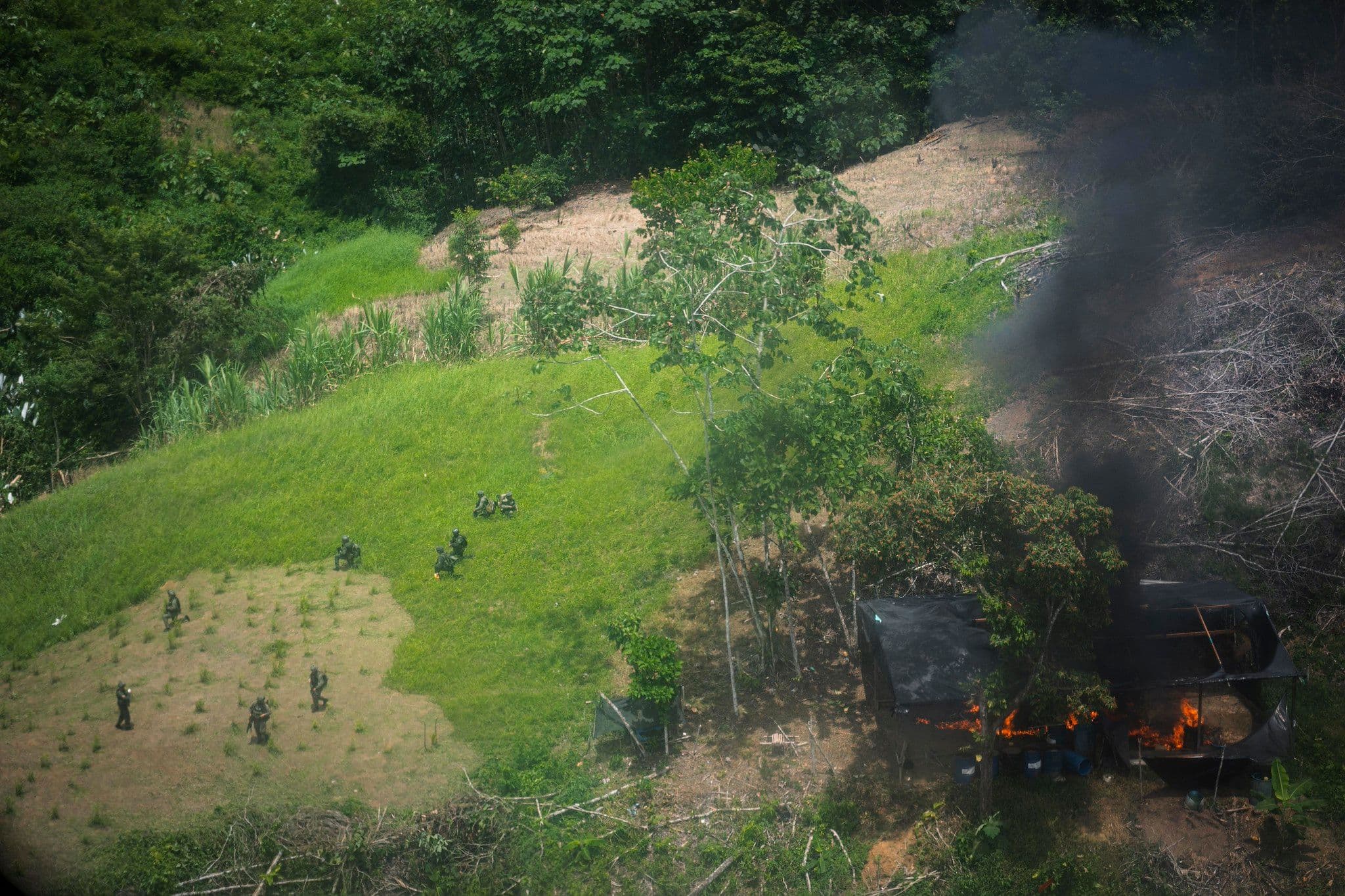 Vista aérea de militares desplegados en una zona rural junto a una estructura en llamas que emite humo negro.