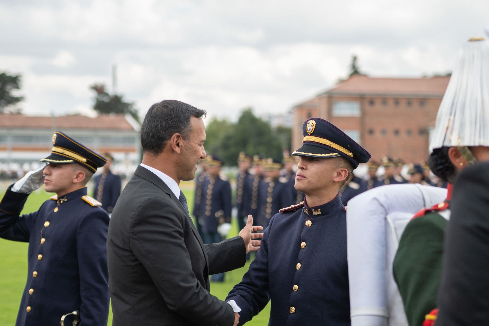 Pedro Sánchez Suárez, de traje, saluda de mano a un cadete en uniforme de gala, durante una ceremonia militar al aire libre con más cadetes formados al fondo.