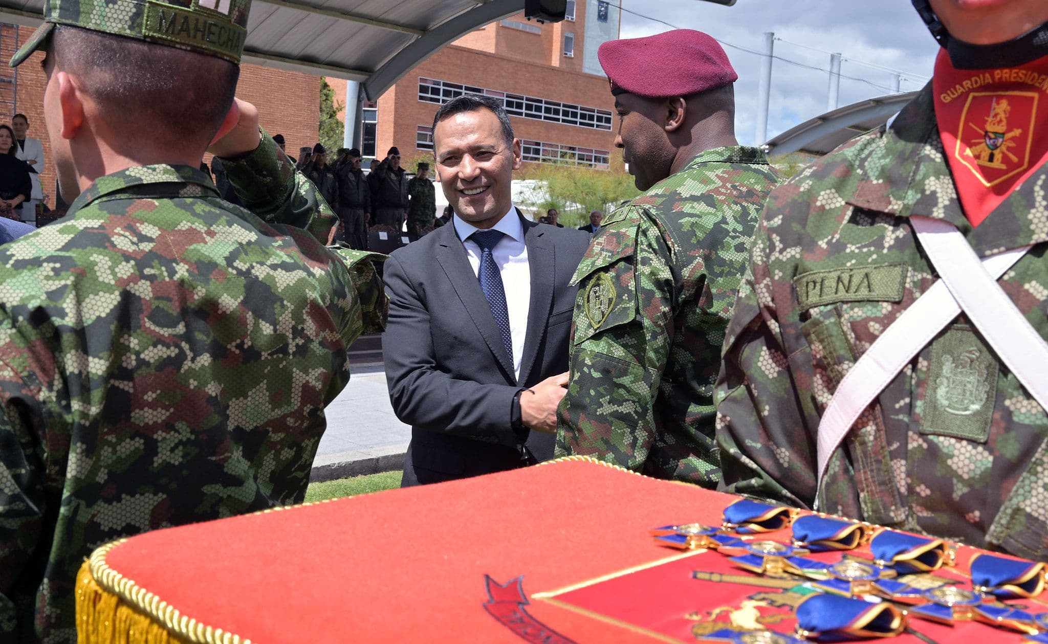 Pedro Sánchez Suárez, de traje, impone una condecoración a un soldado de boina roja, junto a una mesa con medallas y otros militares en formación, durante una ceremonia al aire libre.
