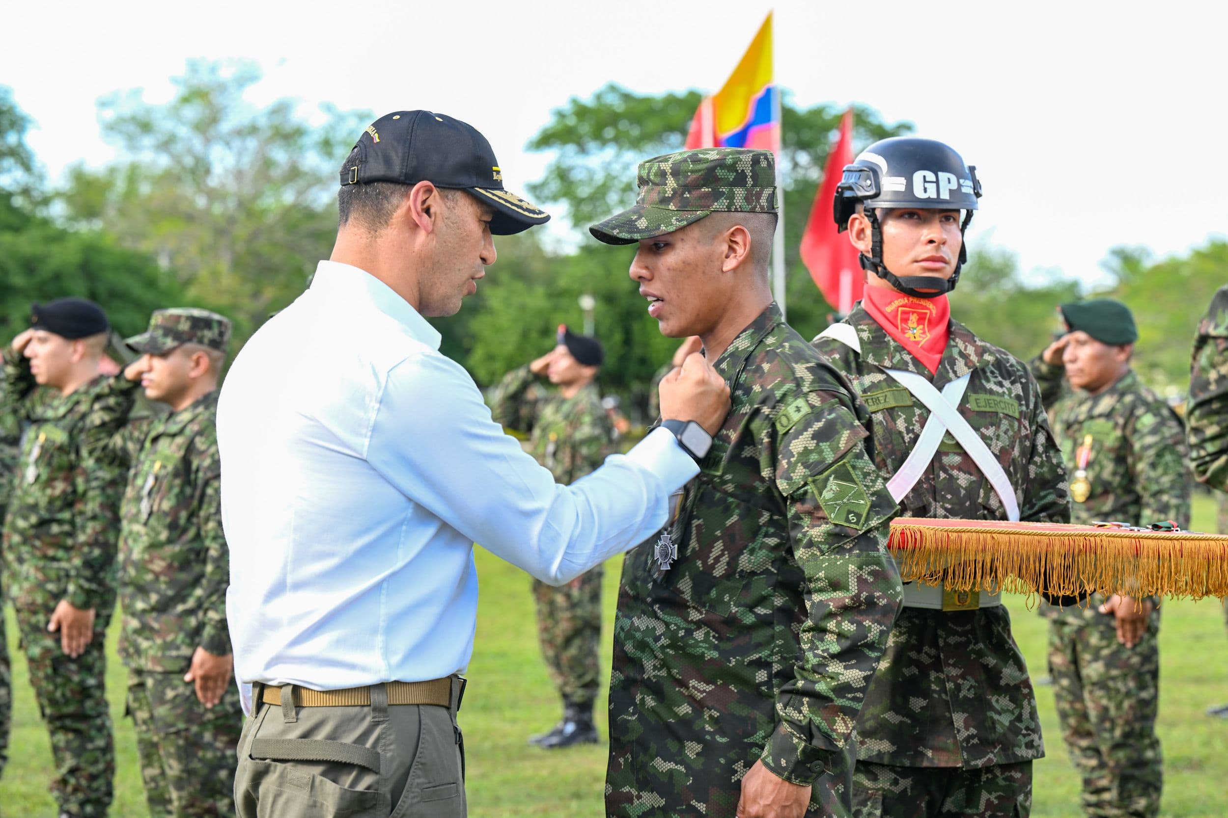 Pedro Sánchez Suárez, de civil, impone una condecoración en el uniforme de un soldado durante una ceremonia militar al aire libre, con banderas y más tropas en formación al fondo.