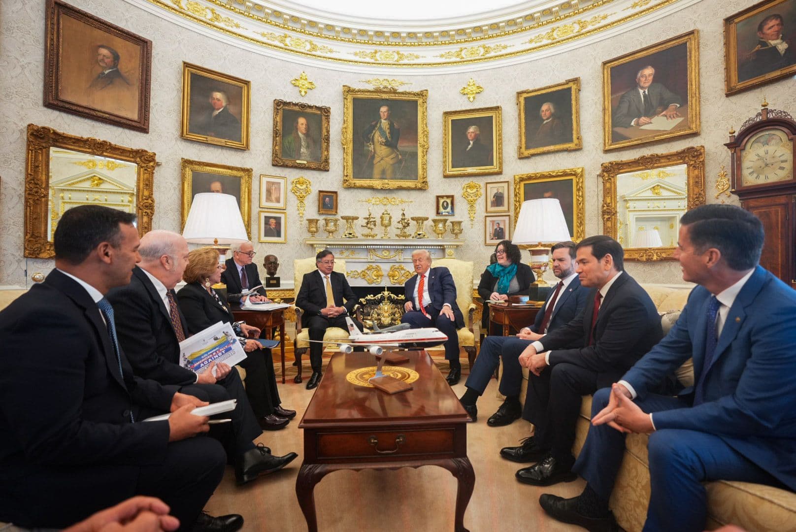 Reunión en la Oficina Oval de la Casa Blanca entre delegaciones de Colombia y Estados Unidos, sentados en sofás alrededor de una mesa con una maqueta de avión presidencial, con retratos históricos en las paredes al fondo.