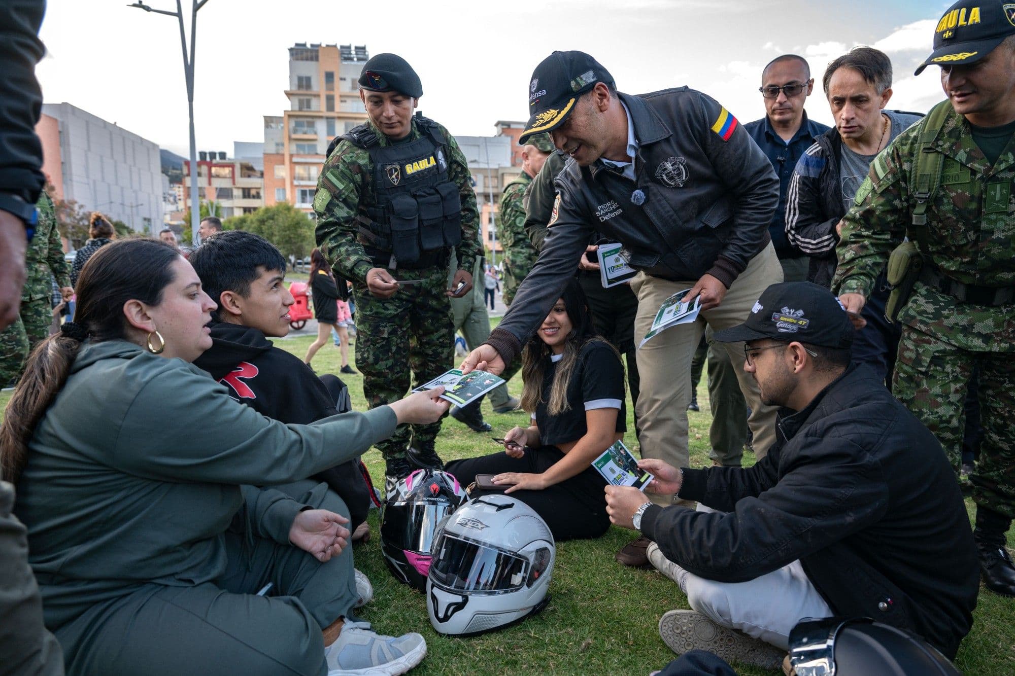 Pedro Sánchez Suárez entrega un volante a un grupo de jóvenes sentados en el pasto, junto a personal del GAULA y otros funcionarios, en un parque urbano.