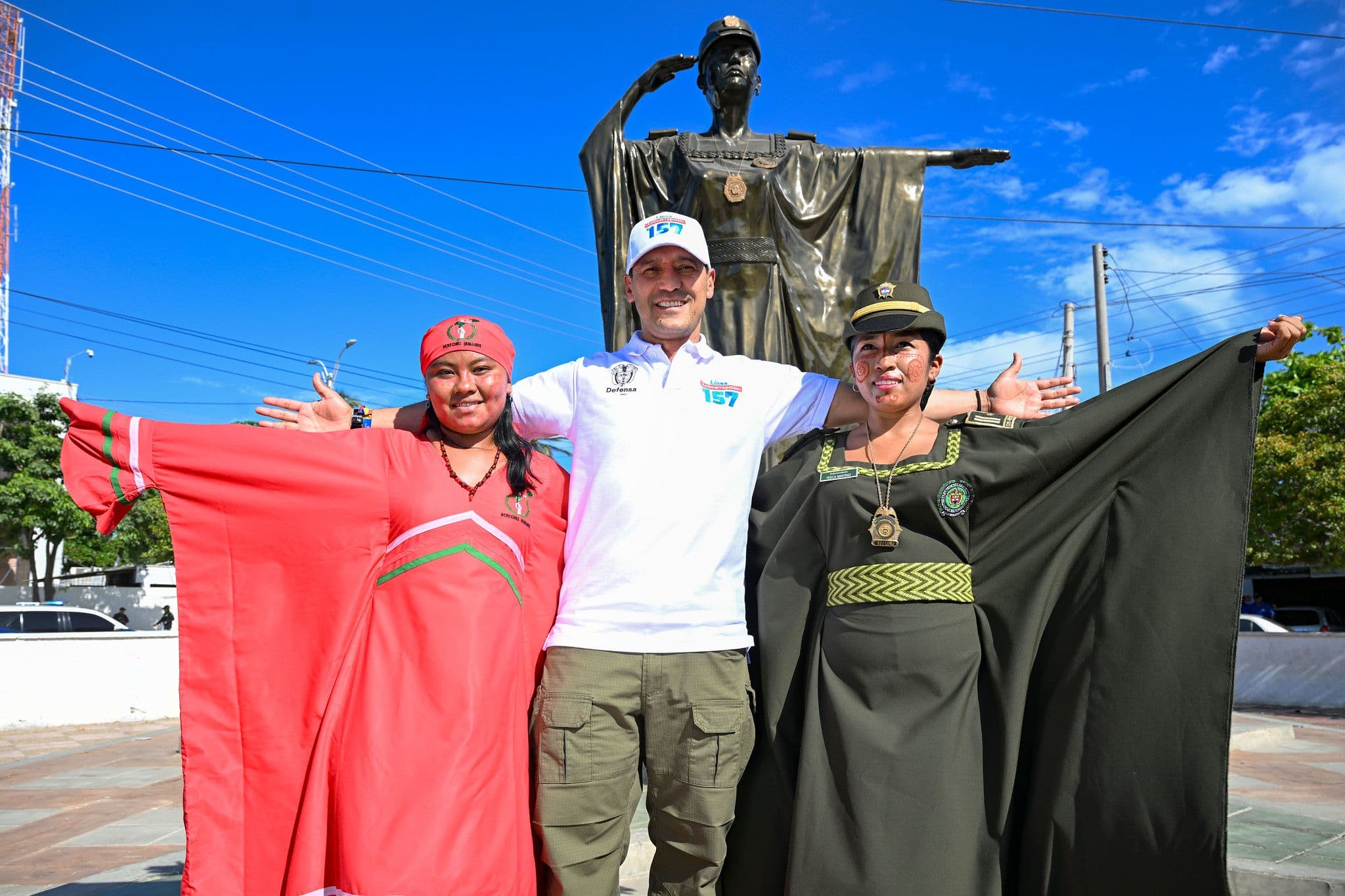 Pedro Sánchez Suárez, con camiseta de la Línea Anticorrupción 157, posa entre dos mujeres que despliegan las capas de un uniforme representado en una estatua de un policía saludando, frente a un monumento al aire libre.