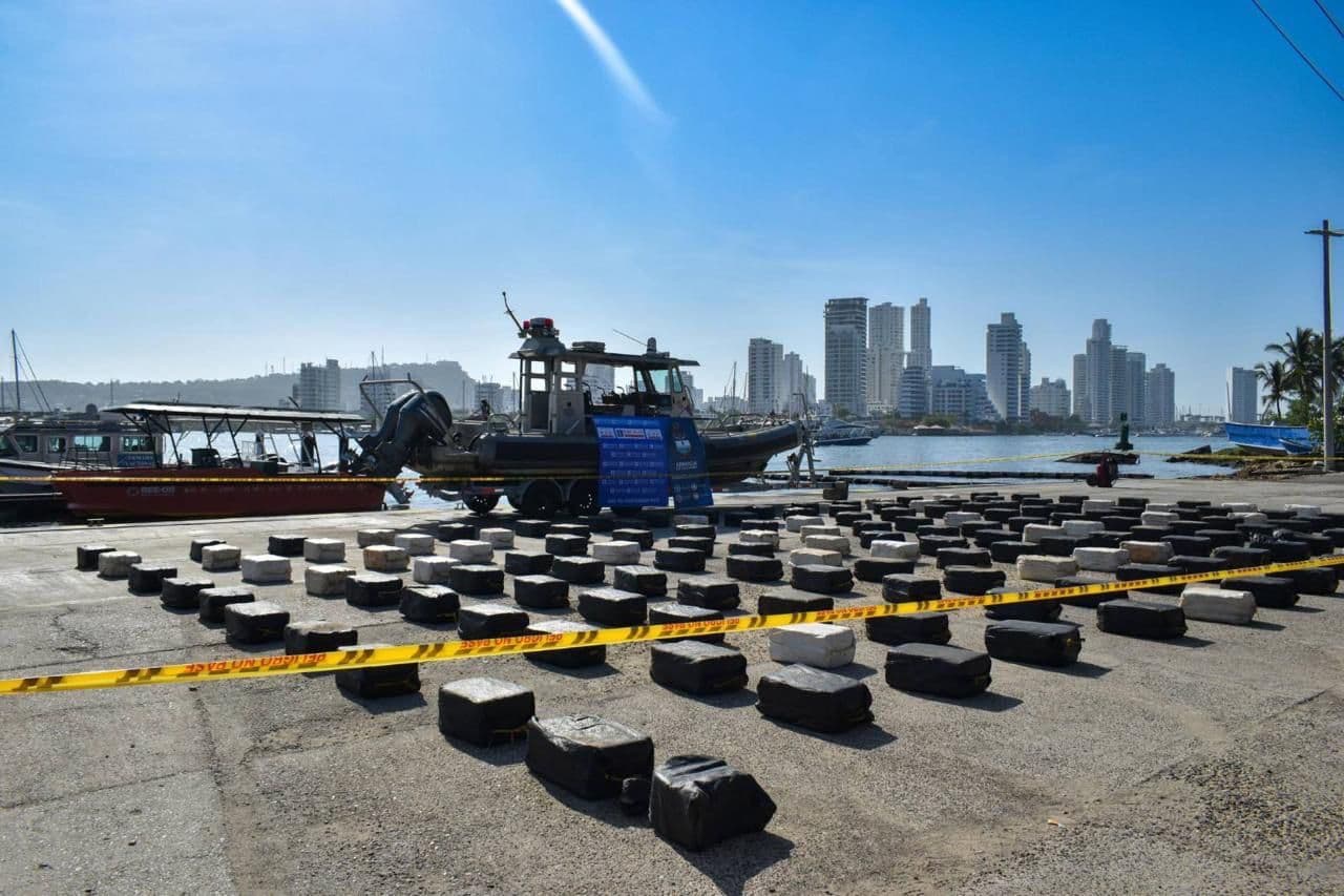 Filas de paquetes envueltos, presuntamente droga incautada, exhibidos en un muelle acordonado con cinta de seguridad, con una lancha de la Armada y el perfil urbano de Cartagena al fondo.