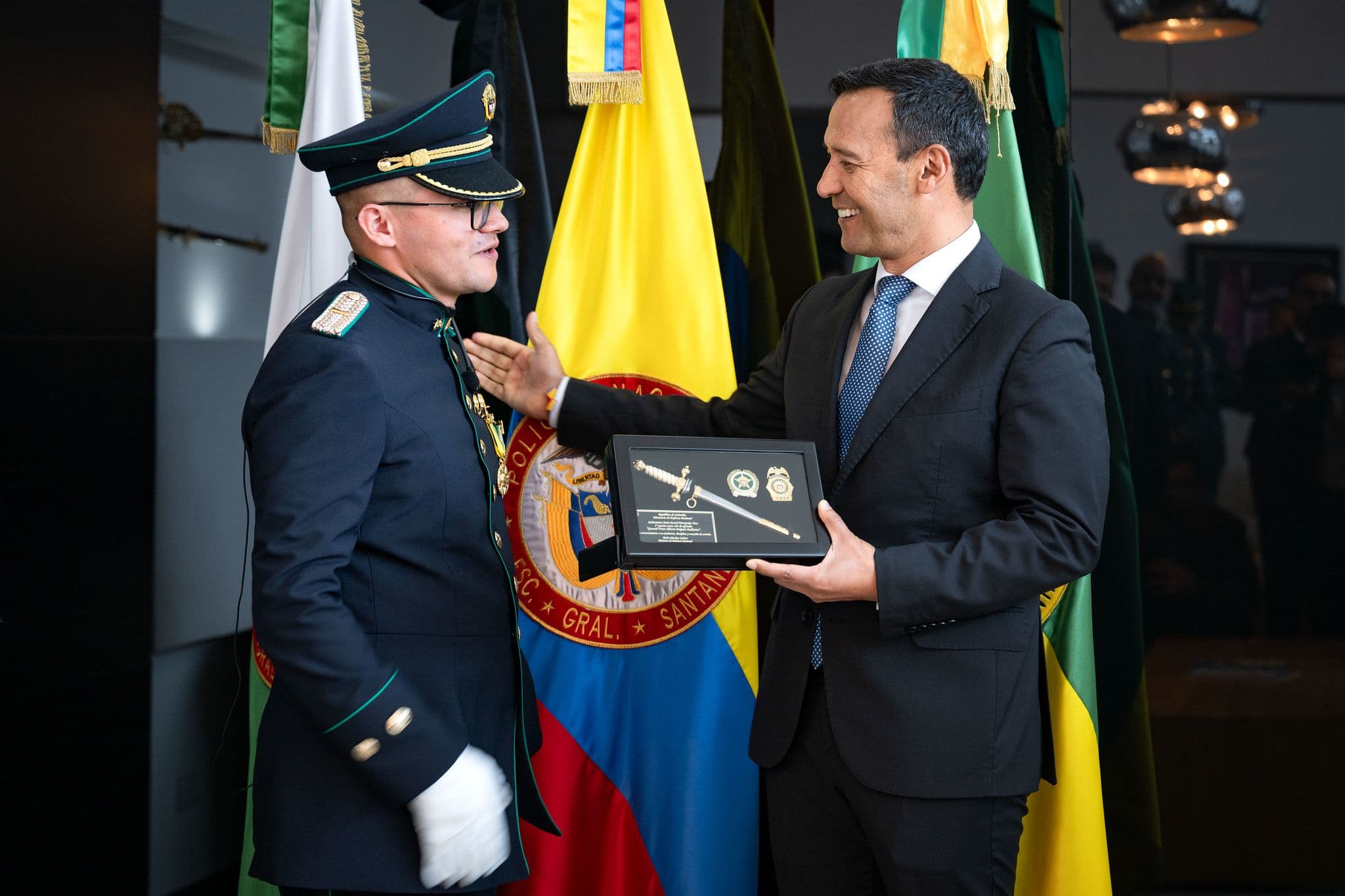 Pedro Sánchez Suárez, de traje, entrega una espada conmemorativa enmarcada a un cadete de policía en uniforme de gala, frente a banderas de Colombia y estandartes institucionales.