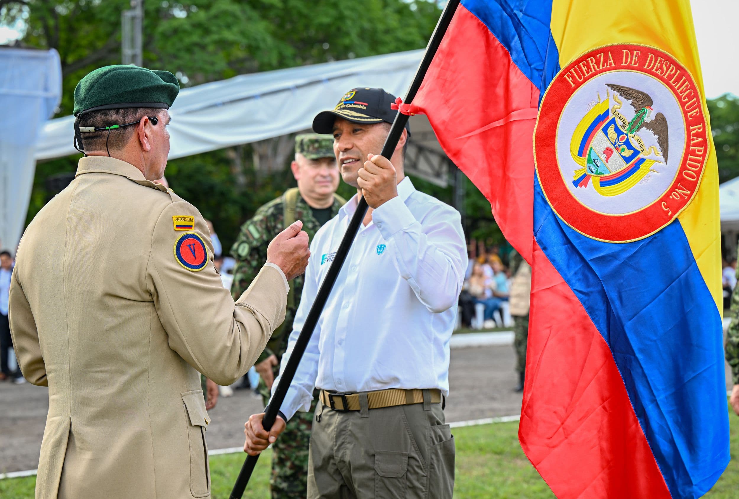 Pedro Sánchez Suárez, vestido de civil, sostiene un asta con la bandera de la Fuerza de Despliegue Rápido No. 5, frente a un oficial en uniforme de gala, durante un acto de activación militar al aire libre.