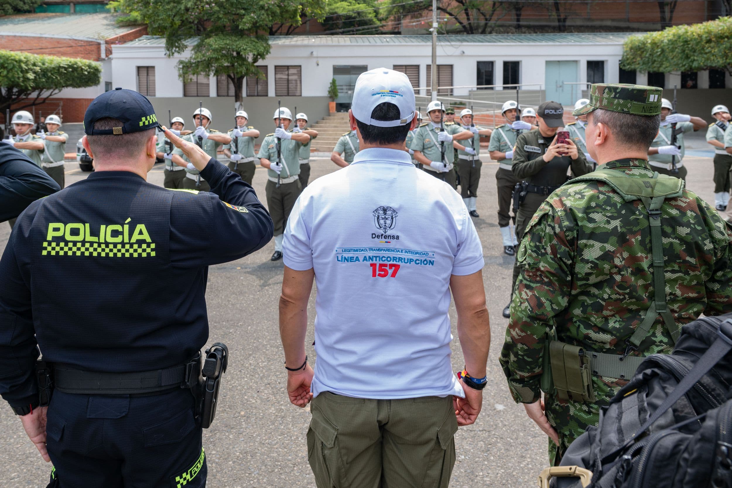 Pedro Sánchez Suárez, de espaldas, con camiseta de la Línea Anticorrupción 157, junto a un policía y un militar, frente a un pelotón de cadetes con casco haciendo el saludo militar en un patio.