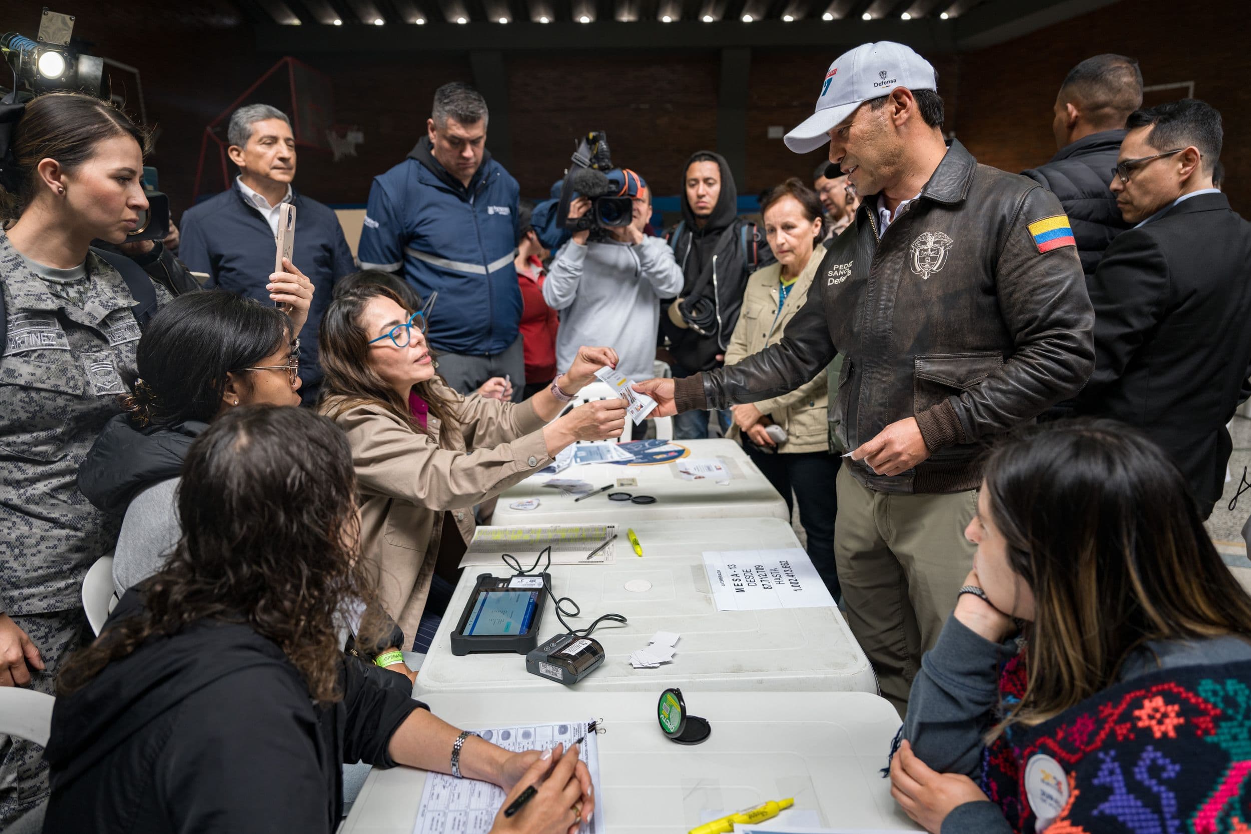 Pedro Sánchez Suárez, de civil, recibe un documento electoral de manos de una jurado de votación en una mesa de sufragio, rodeado de personal militar, prensa y otras personas, en un puesto de votación.