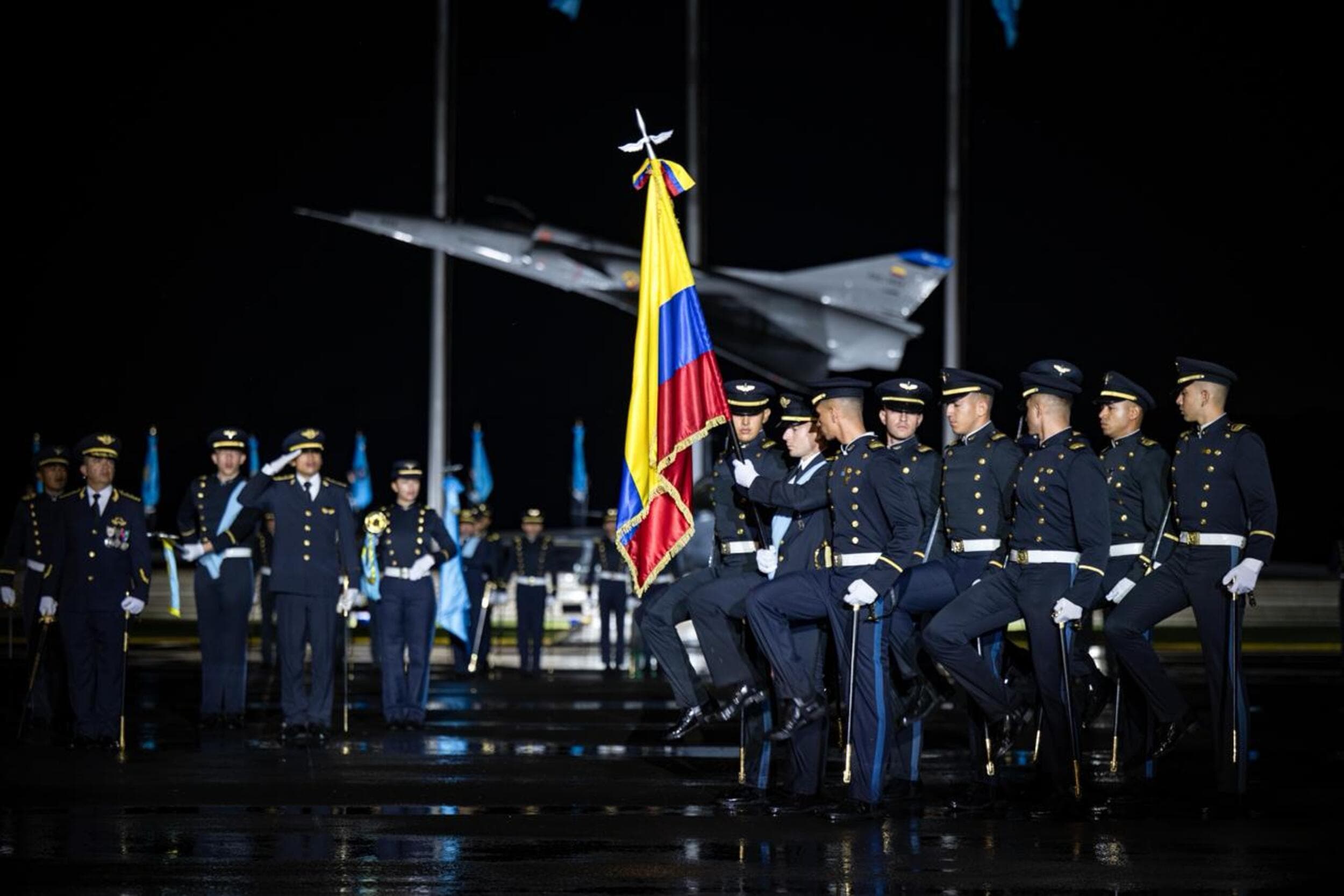 Bandera de Colombia en primer plano sostenida por un oficial durante una ceremonia militar nocturna, con miembros de la Fuerza Aérea en uniforme de gala y un avión de combate al fondo.