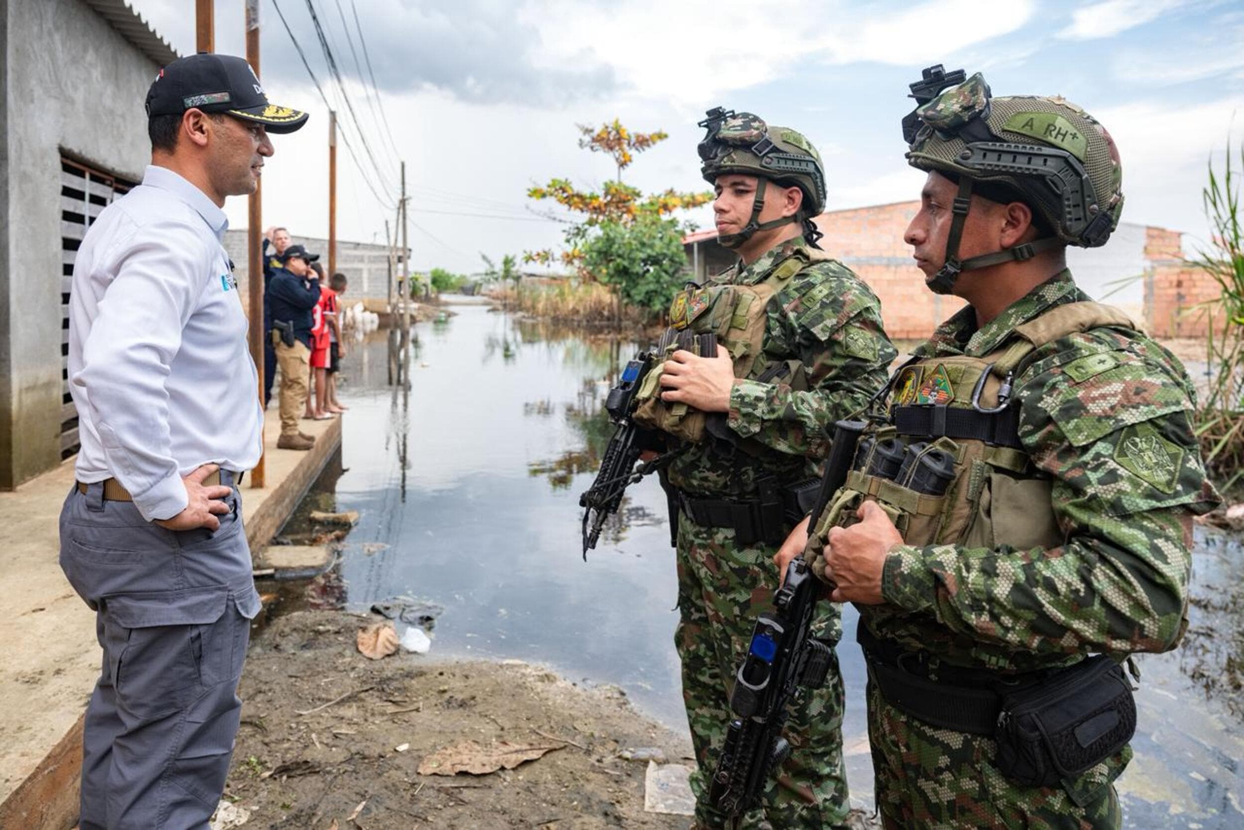 Pedro Sánchez Suárez, de civil, conversa con dos militares armados y en equipo táctico completo junto a una calle inundada, con niños y más personas observando al fondo.