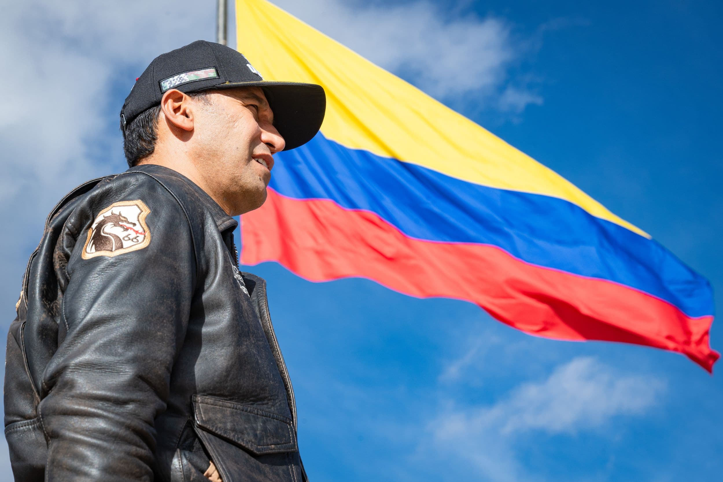 Pedro Sánchez Suárez, de perfil, con gorra y chaqueta de cuero con un parche de escudo, frente a la bandera de Colombia ondeando contra un cielo azul con nubes.